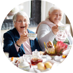 Two elderly women smiling and raising tea cups at a table with tea and pastries.