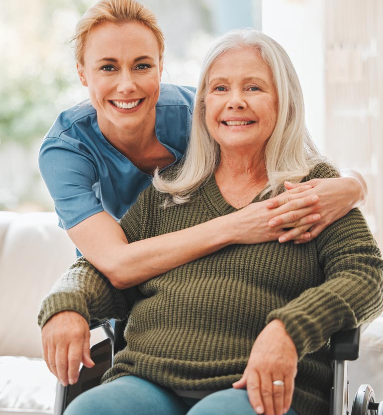 Smiling caregiver in blue scrubs embracing a senior woman seated in a wheelchair indoors.