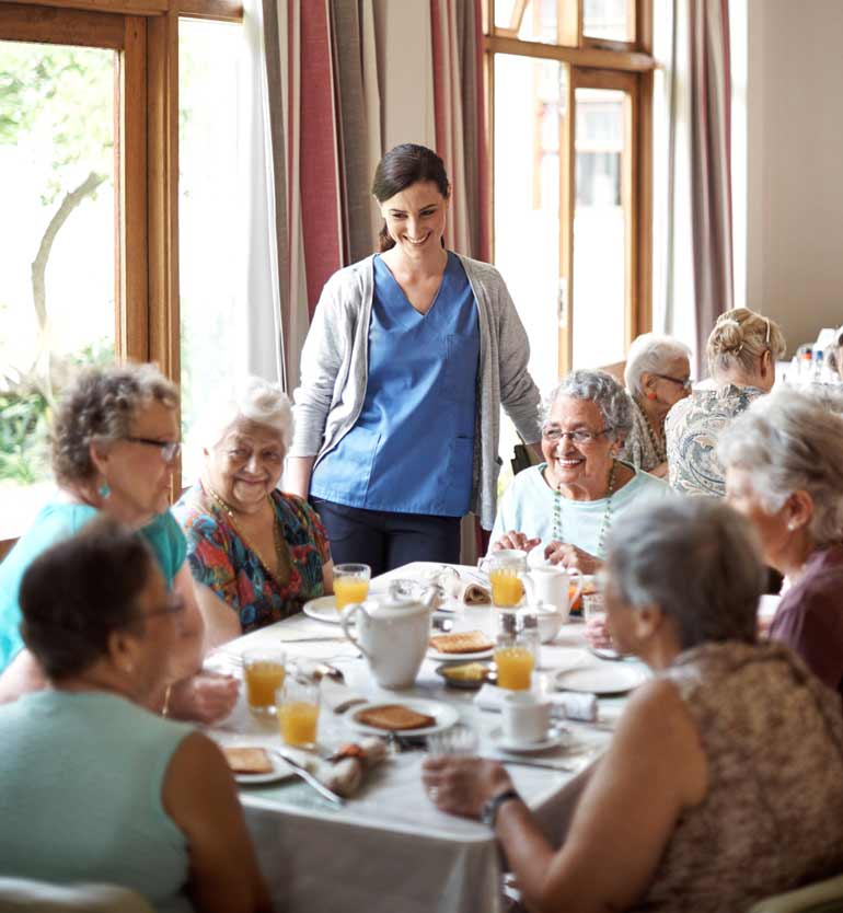 A caregiver in blue scrubs and seniors enjoy breakfast together at a well-lit senior living facility.