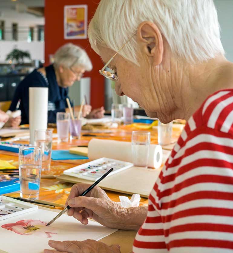 Elderly woman with short white hair in striped shirt painting a flower at an art class table.