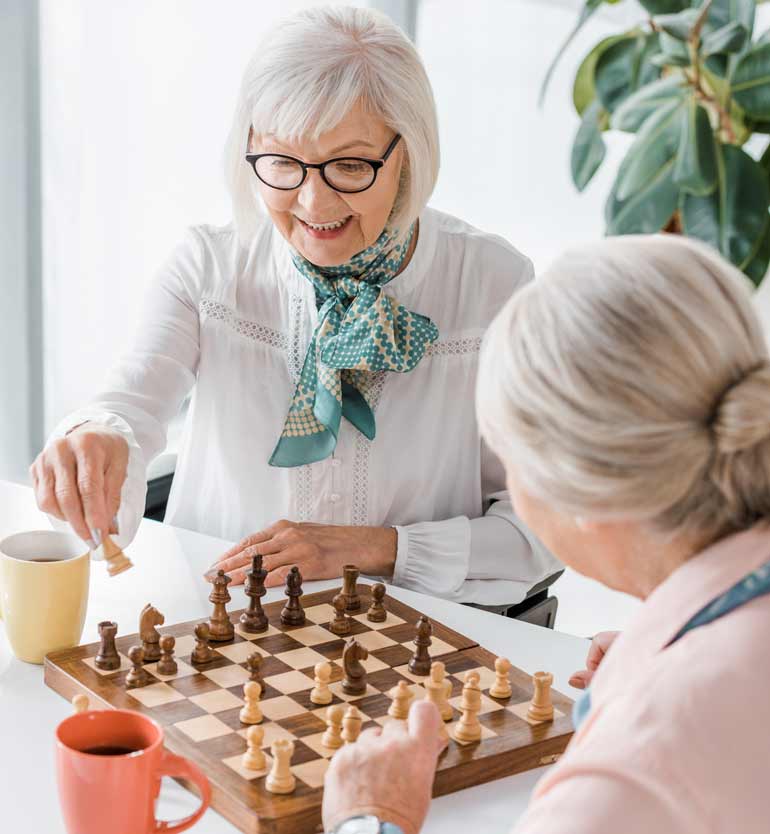 Two elderly women playing chess at a table, one making a move with coffee cups beside them.