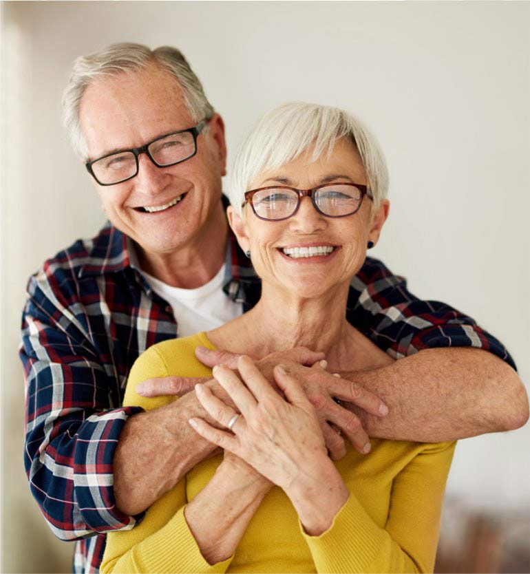Smiling senior couple, the man hugging the woman from behind in a brightly lit room.