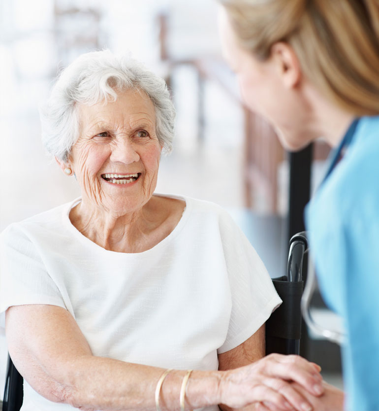 Elderly woman smiling warmly at a caregiver who is holding her hand in a supportive environment.