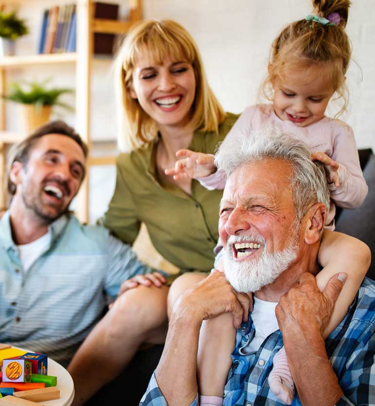 Senior man laughing joyfully while holding his young granddaughter on his shoulders, surrounded by family.