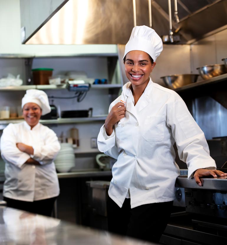 Two female chefs smiling and standing in a professional kitchen with stainless steel appliances.