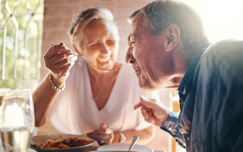 Two senior people enjoying a meal together, one feeding the other with a spoon.