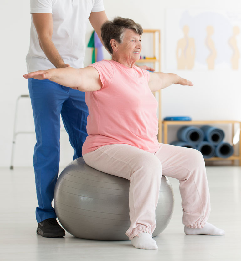 Senior woman doing physical therapy exercises with a therapists help on a stability ball.