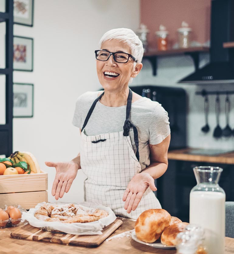 Smiling woman with short white hair and glasses presenting freshly baked bread in her kitchen.