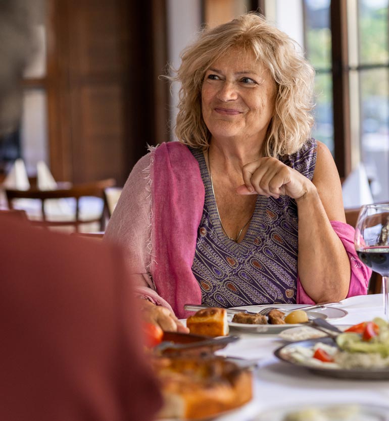 Senior woman enjoying a meal with friends at a restaurant, smiling and wearing a patterned top.