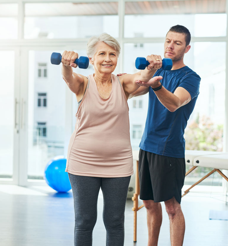 Senior woman lifting dumbbells with assistance from a trainer in a bright, modern gym.