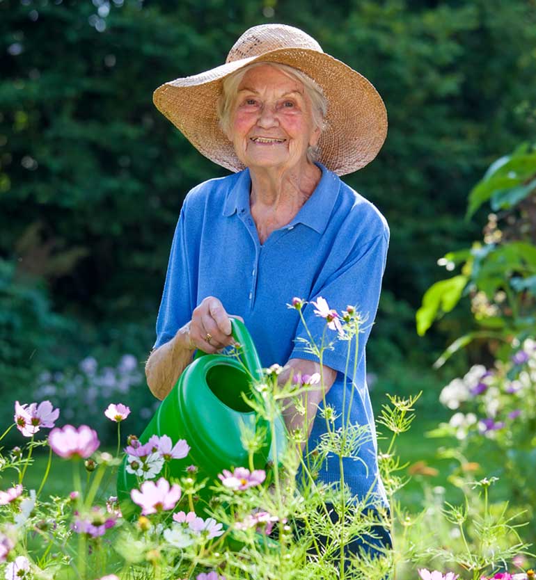 Elderly woman wearing a sun hat and blue shirt, watering flowers in a garden with a green watering can.