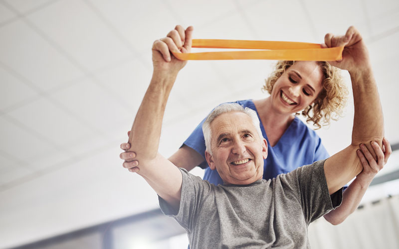 Elderly man exercising with a resistance band assisted by a female caregiver in a bright room.